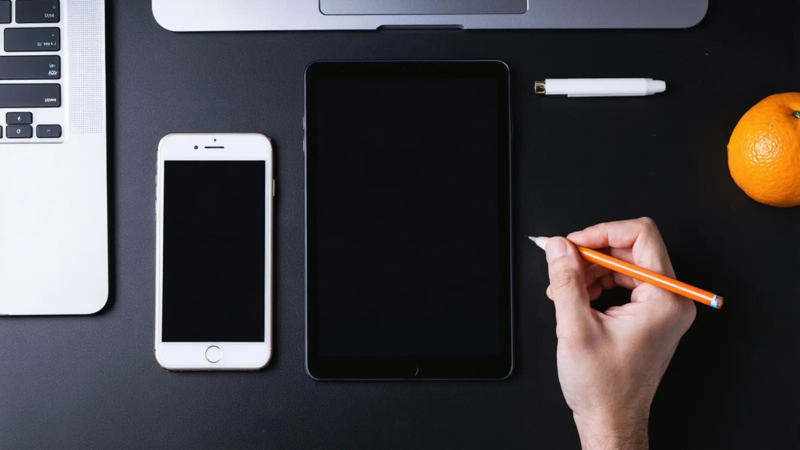 A phone, a tablet and a laptop on a dark desk, with a hand holding an orange pencil