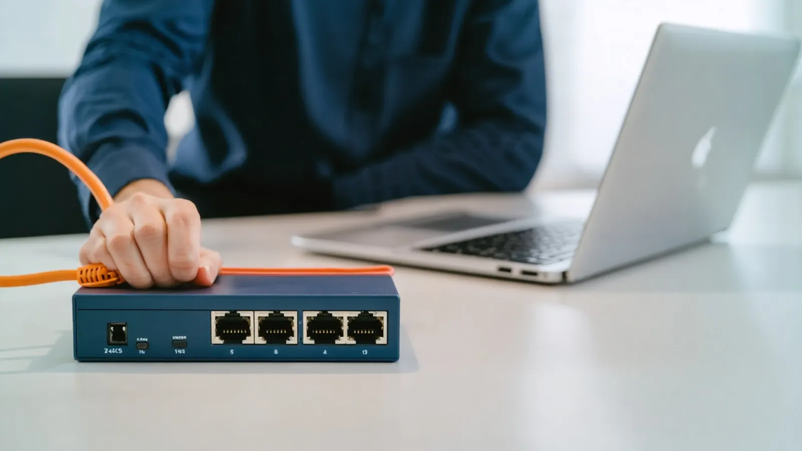 A hand plugging an orange network cable into a small switch beside a laptop